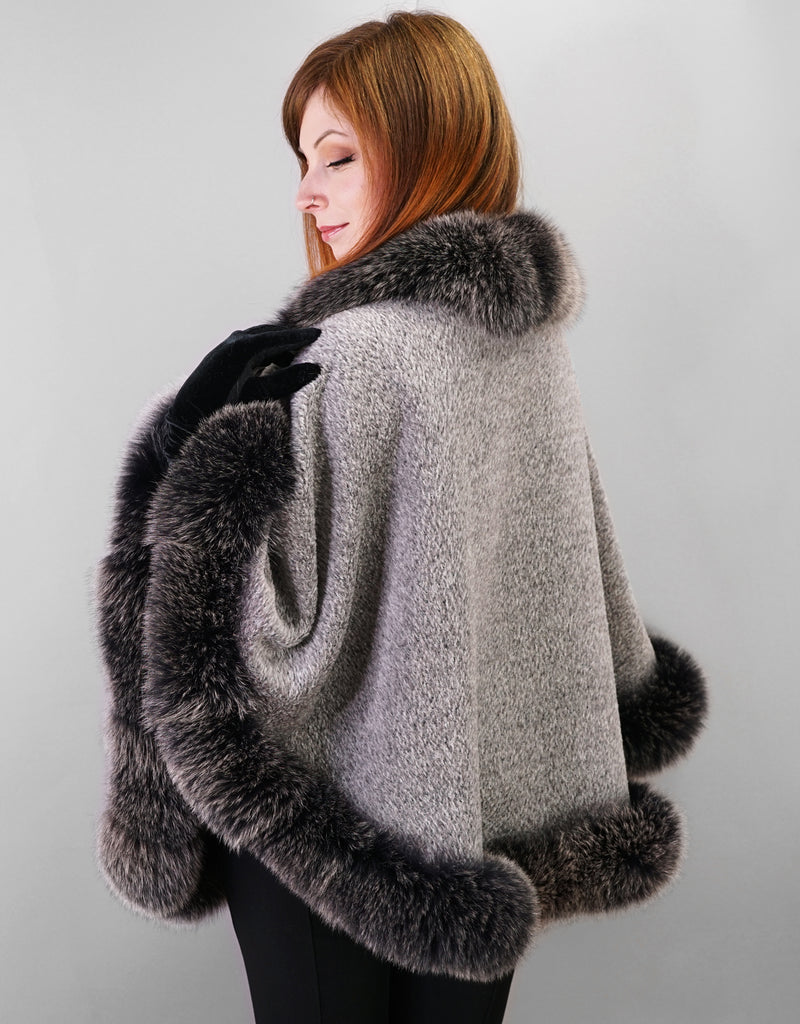Back view: woman wearing silver alpaca wool cape with black fox trim, the black fox has white tips and is called black snow