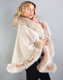 A blonde woman with green eyes is wearing a medium length beige cape with blush fox trim. Medium length reaches mid thigh to knee. The cape has velvetty looking woven Alpaca wool with fluffy shiny fox trim.