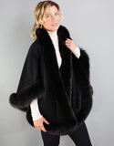A blonde woman with green eyes is wearing a medium length black cape. Medium length reaches mid thigh to knee.  The cape has velvetty looking black woven Alpaca wool with fluffy shiny black fox trim.