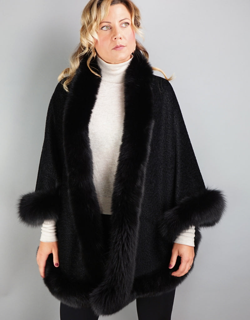 A blonde woman with green eyes is wearing a medium length black cape. Medium length reaches mid thigh to knee. The cape has velvetty looking black woven Alpaca wool with fluffy shiny black fox trim.
