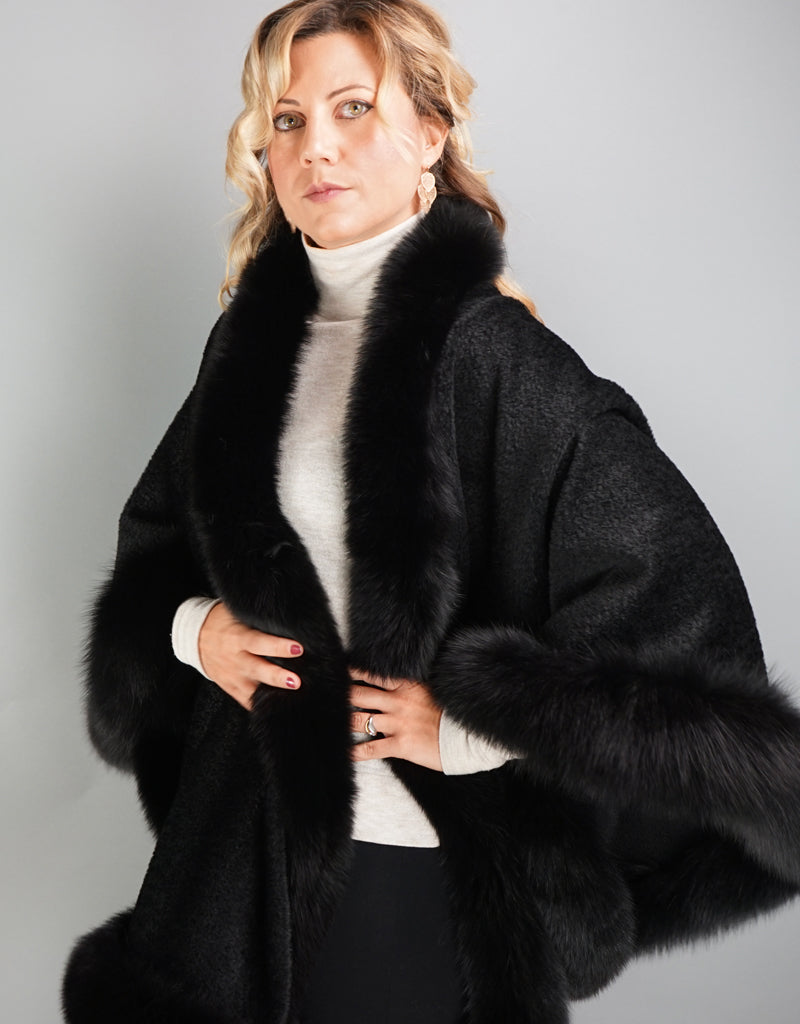 A blonde woman with green eyes is wearing a medium length black cape. Medium length reaches mid thigh to knee. The cape has velvetty looking black woven Alpaca wool with fluffy shiny black fox trim