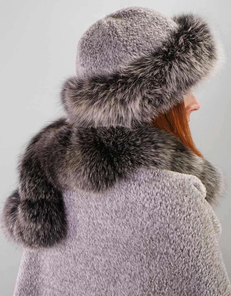 Back view : A woman with red hair is wearing a silver alpaca wool cape with black snow colour fox trim. She is wearing a matching hat with a silver alpaca wool crown and black snow fox trim.