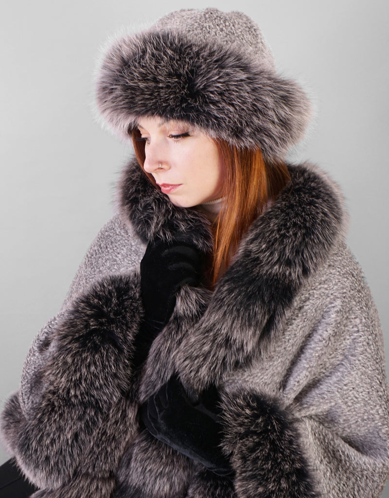 Front view: A woman with red hair is wearing a silver alpaca wool cape with black snow colour fox trim. She is wearing a matching hat with a silver alpaca wool crown and black snow fox trim.