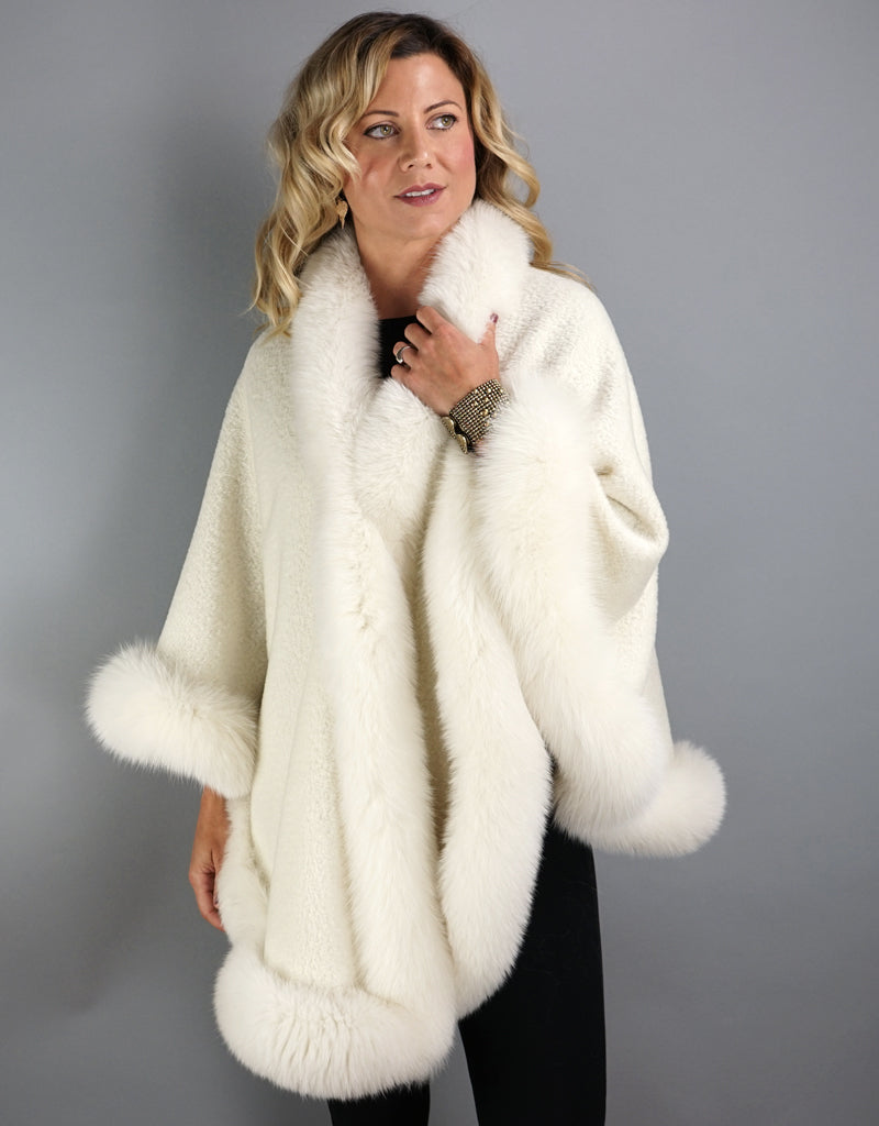 A blonde woman with green eyes is wearing a medium length off white cape. Medium length reaches mid thigh to knee. The cape has velvetty looking woven Alpaca wool with fluffy shiny fox trim