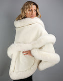 A blonde woman with green eyes is wearing a medium length off white cape. Medium length reaches mid thigh to knee. The cape has velvetty looking woven Alpaca wool with fluffy shiny fox trim.